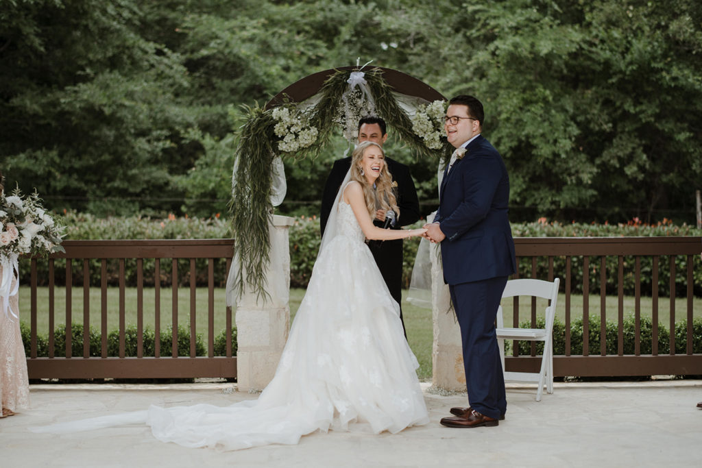blonde bride makes her tall, dark and handsome groom laugh during their vows with a forest of greenery behind them