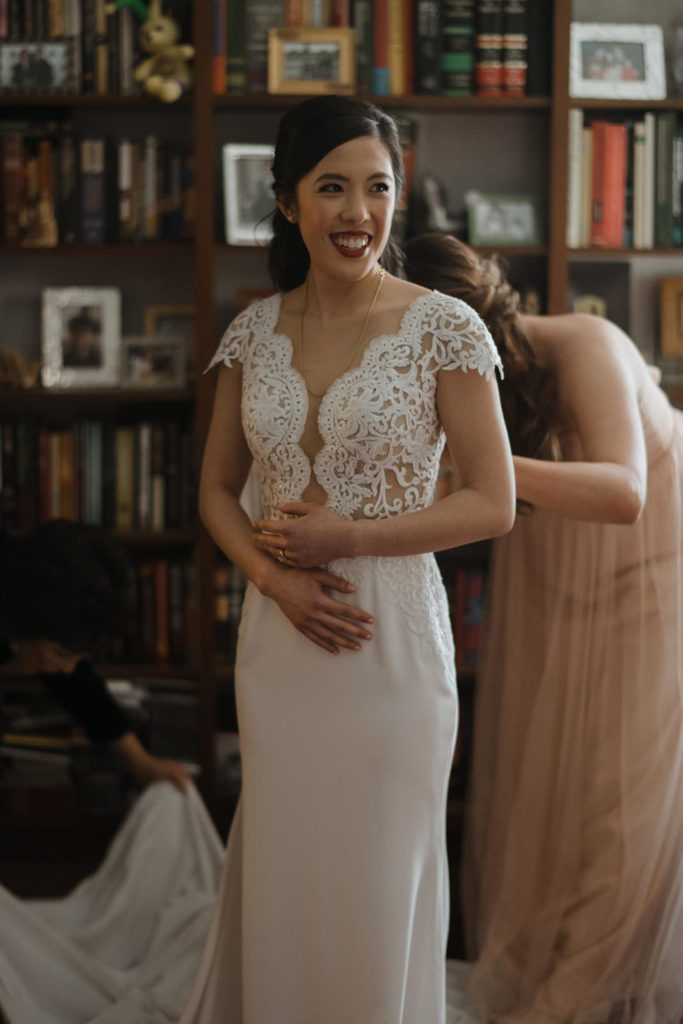 bride gets zipped into her dress by her Maid of Honor on her wedding day in a circular room full of large windows while she looks in the mirror