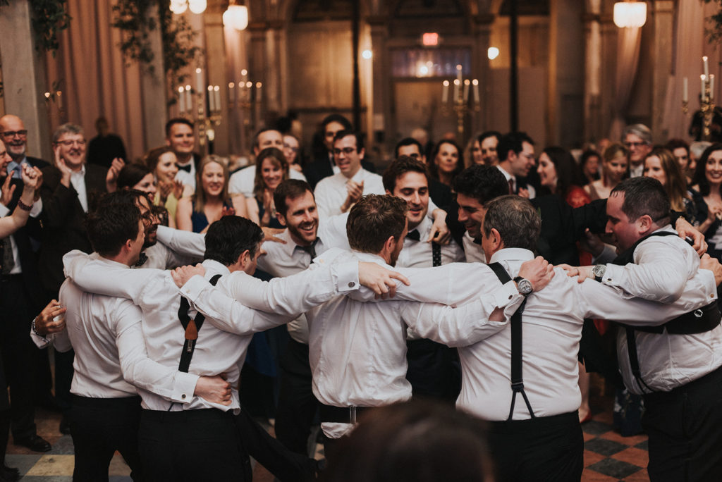 all the groomsmen at a wedding reception form a hugging circle as they dance together