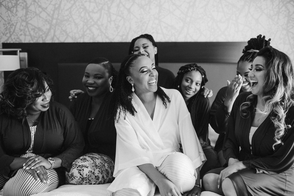 a negro bride and her bridesmaids sit on a bed laughing hysterically as they engage with the photographer
