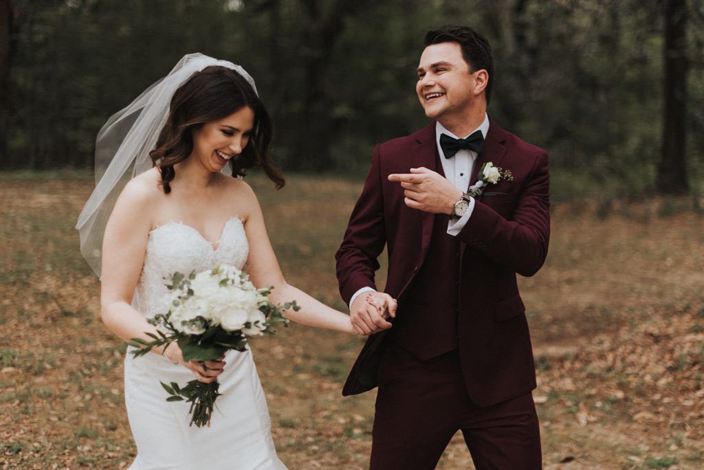 bride carrying a white bouquet and groom wearing a burgundy tux laugh while walking hand in hand through the forest on their wedding day in New Orleans. PHotos by Erin & Geoffrey Photography.