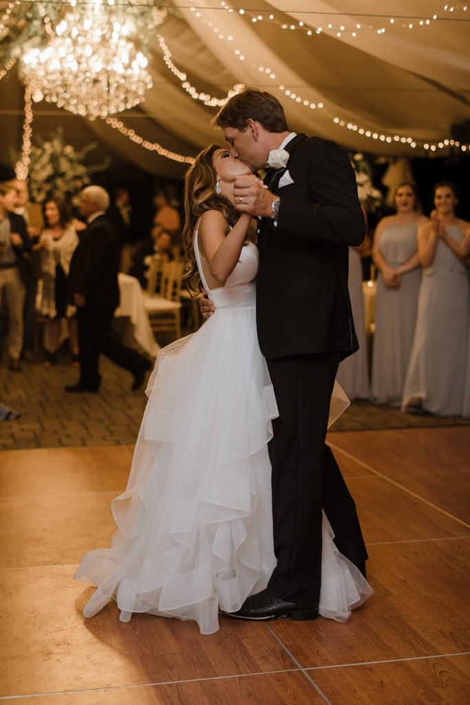 bride and groom kiss during their first dance on a wooden dancefloor under a tent with a chandelier hanging from the ceiling with their wedding party and guests watching