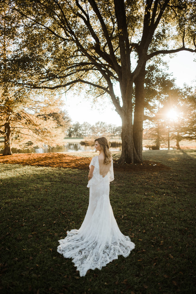 gorgeous sunset shot of a bride in New Orleans standing on a grassy lawn with a pond behind her and the sun filtering through the leaves of the trees beside her